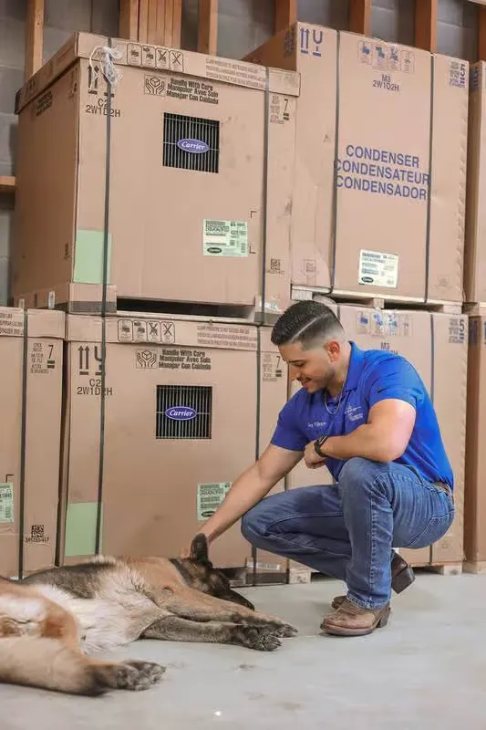 Frontier technician crouching to pet a German Shepherd beside stacked Carrier condenser boxes