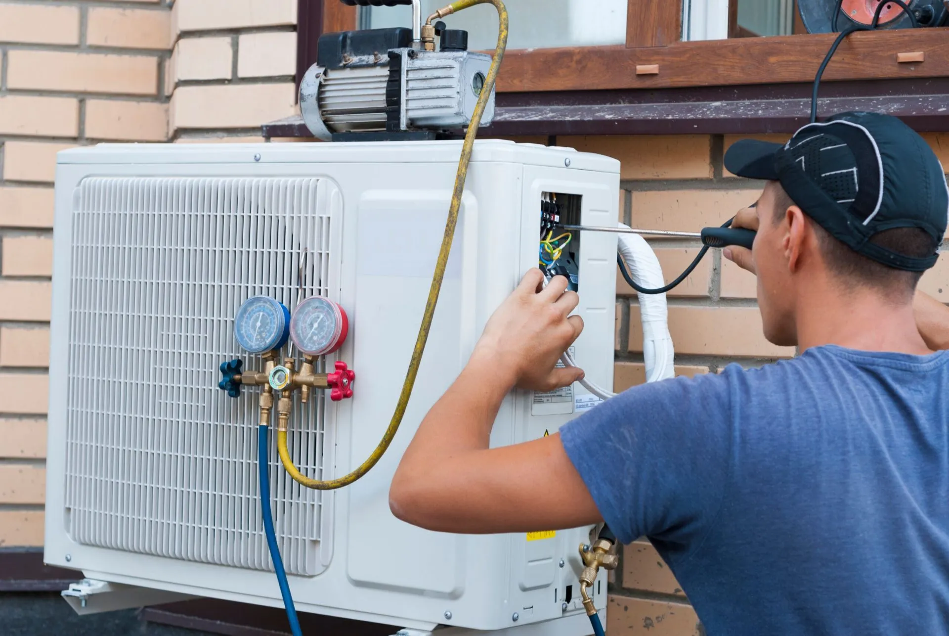 A technician in a blue shirt works on an outdoor air conditioning unit connected to a vacuum pump and pressure gauges.