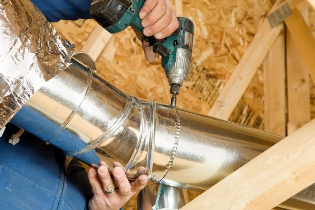 Person using a drill to install HVAC ductwork in an attic, wooden beams visible.