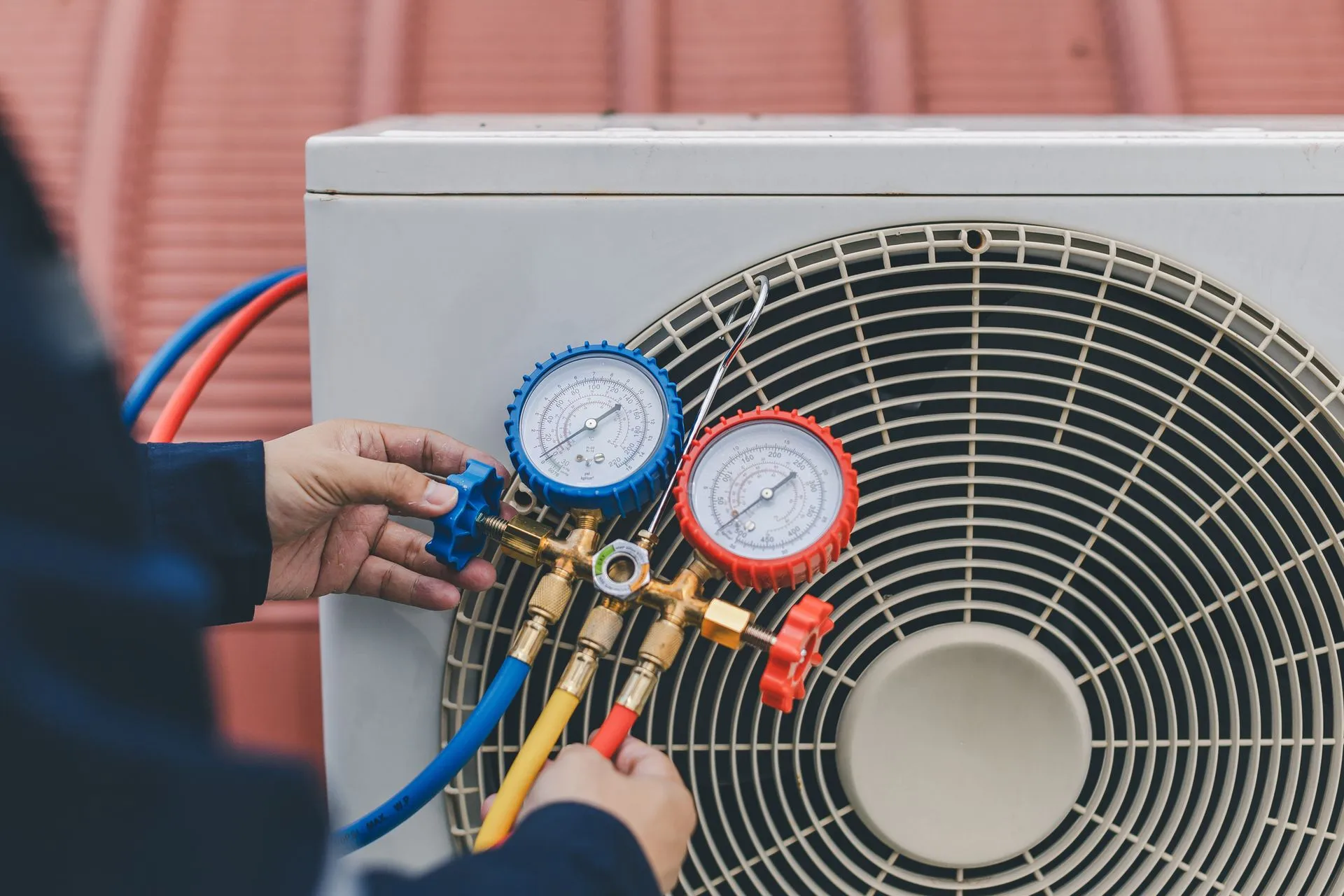 A person connects gauges to an outdoor AC unit. Blue and red hoses and gauges are attached.