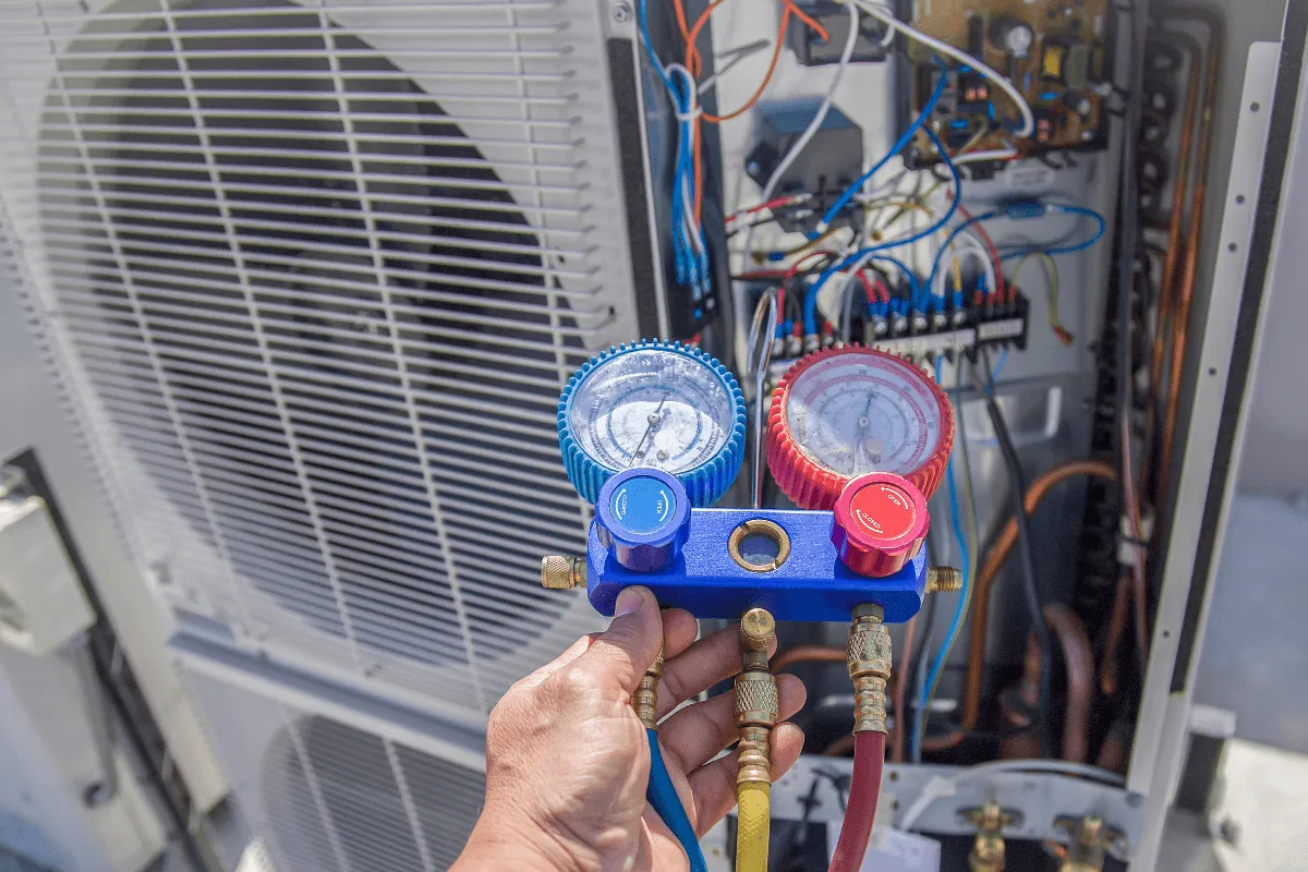 Hand holding HVAC manifold gauges in front of an outdoor air conditioner unit.