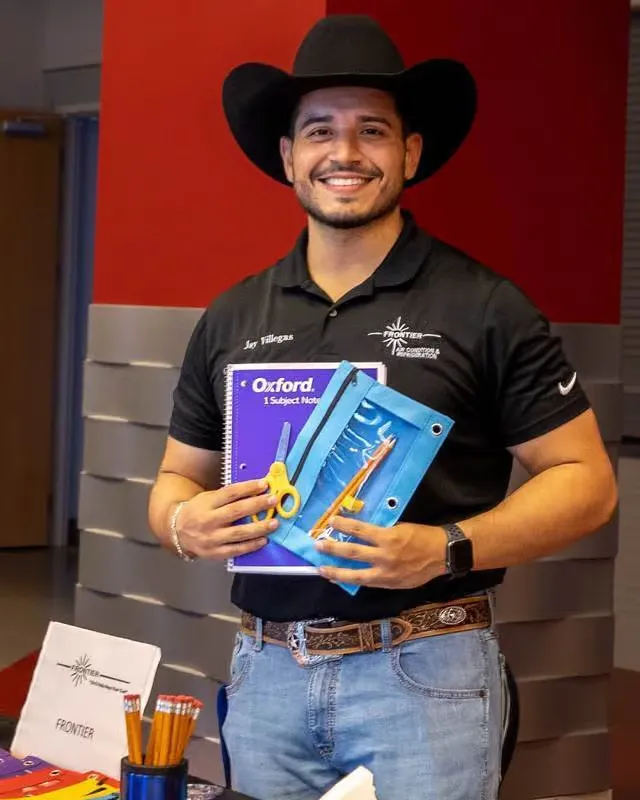 Jay Villegas in a cowboy hat holding notebooks and pencils at a school supply drive