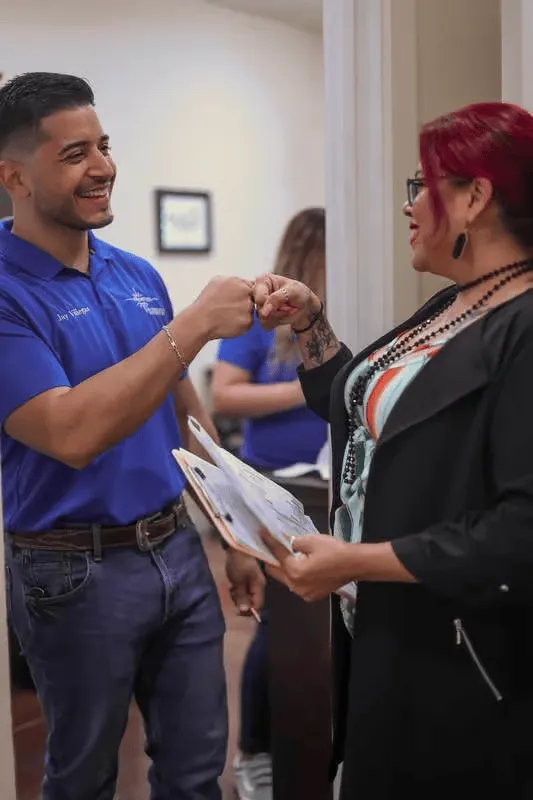 Two people greet each other with a fist bump in a hallway, one holding papers.