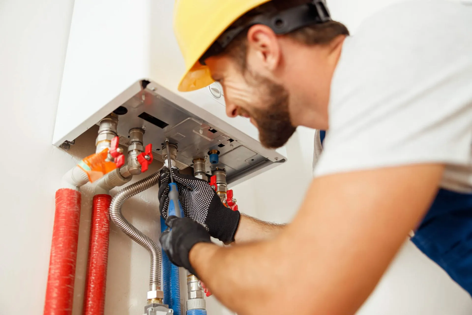 A technician in a yellow hard hat repairs a boiler, using tools, indoors.