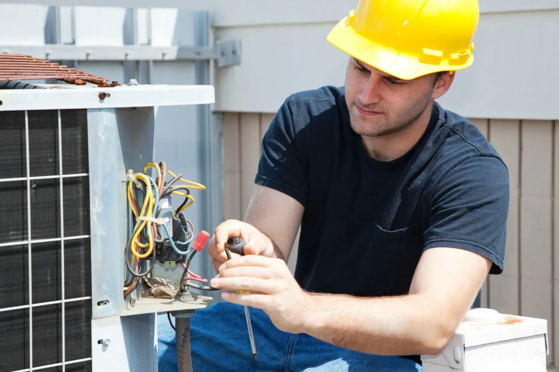 Man in a hard hat repairing an air conditioning unit outside, using tools on the wires.